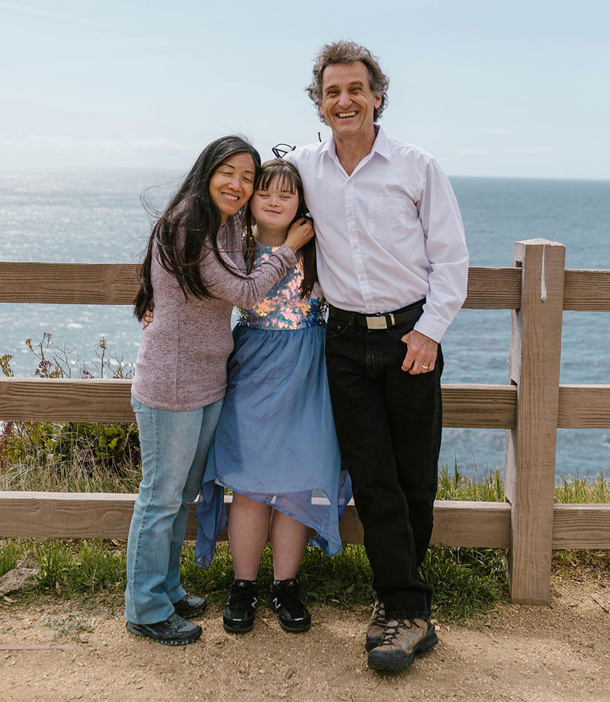 parents hug their daughter with down syndrome while leaning on a fence overlooking the ocean