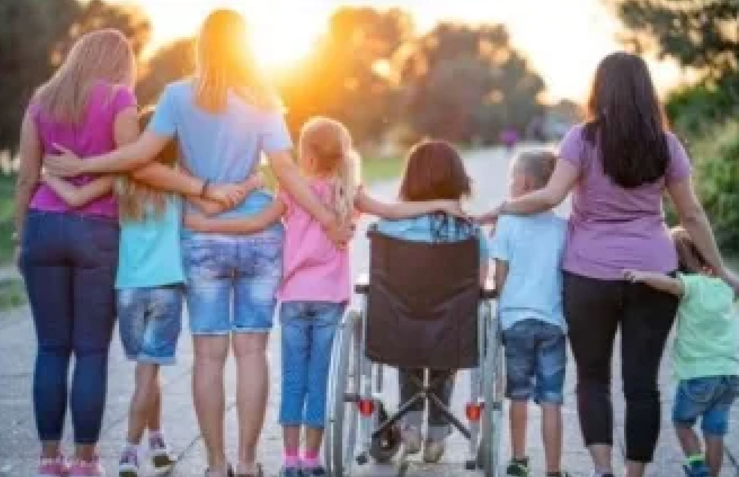 Group of children & adults with and without visual disabilities standing in a line with their backs facing the viewer, arms clasped behind their bodies