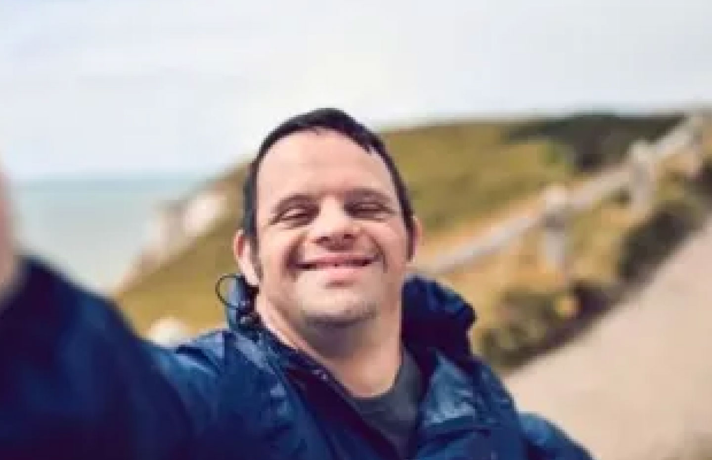 Man with down syndrome taking a selfie on a walking path overlooking a cliff & beach in the blurry background