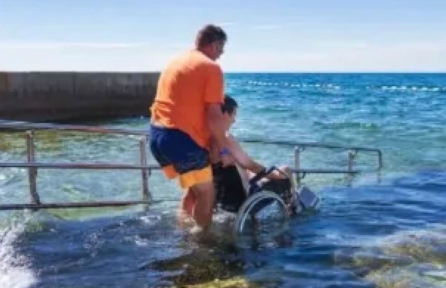 Man using a wheelchair being assisted down a ramp into the ocean by another man without visual disabilities