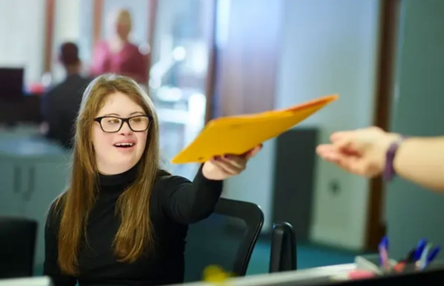 Woman with down syndrome, long brown hair, black turtleneck shirt, & black rimmed glasses handing a folder to someone at work