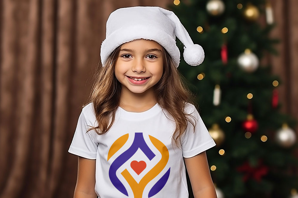 little girl wearing a white santa hat & white always you signature logo t-shirt in front of a christmas tree & brown curtain background