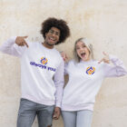 2 young adults pointing to their white sweatshirts with Always You graphics & logos with excited smiles in front of a cream-colored cement background