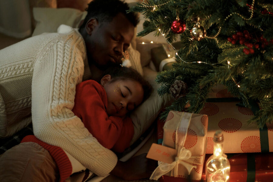 a father & young boy sleep cuddled together on the floor under their christmas tree