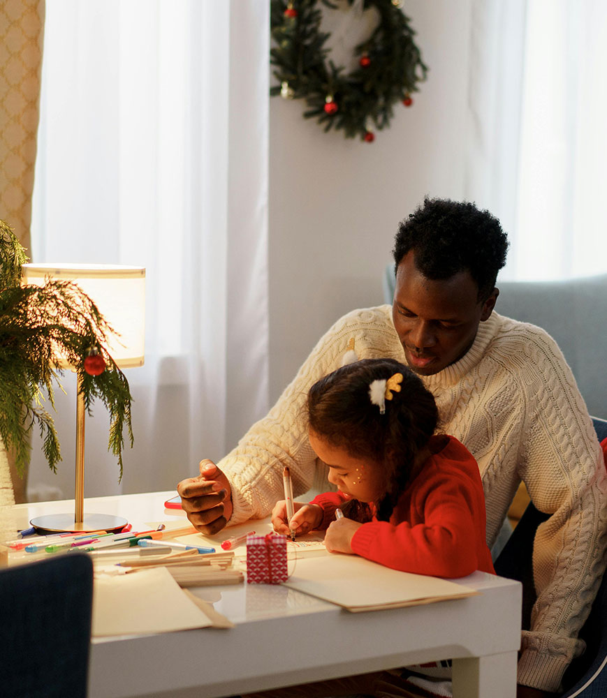 father & daughter sit at a small table writing a letter or crafting together with soft lighting & christmas decorations in the background