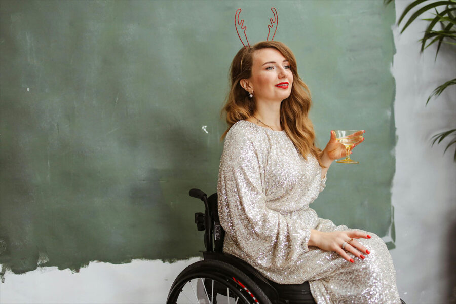 a young woman in a wheelchair is wearing a sparkly silver dress & reindeer antlers & holding a cocktail in front of a grey & sage colored wall