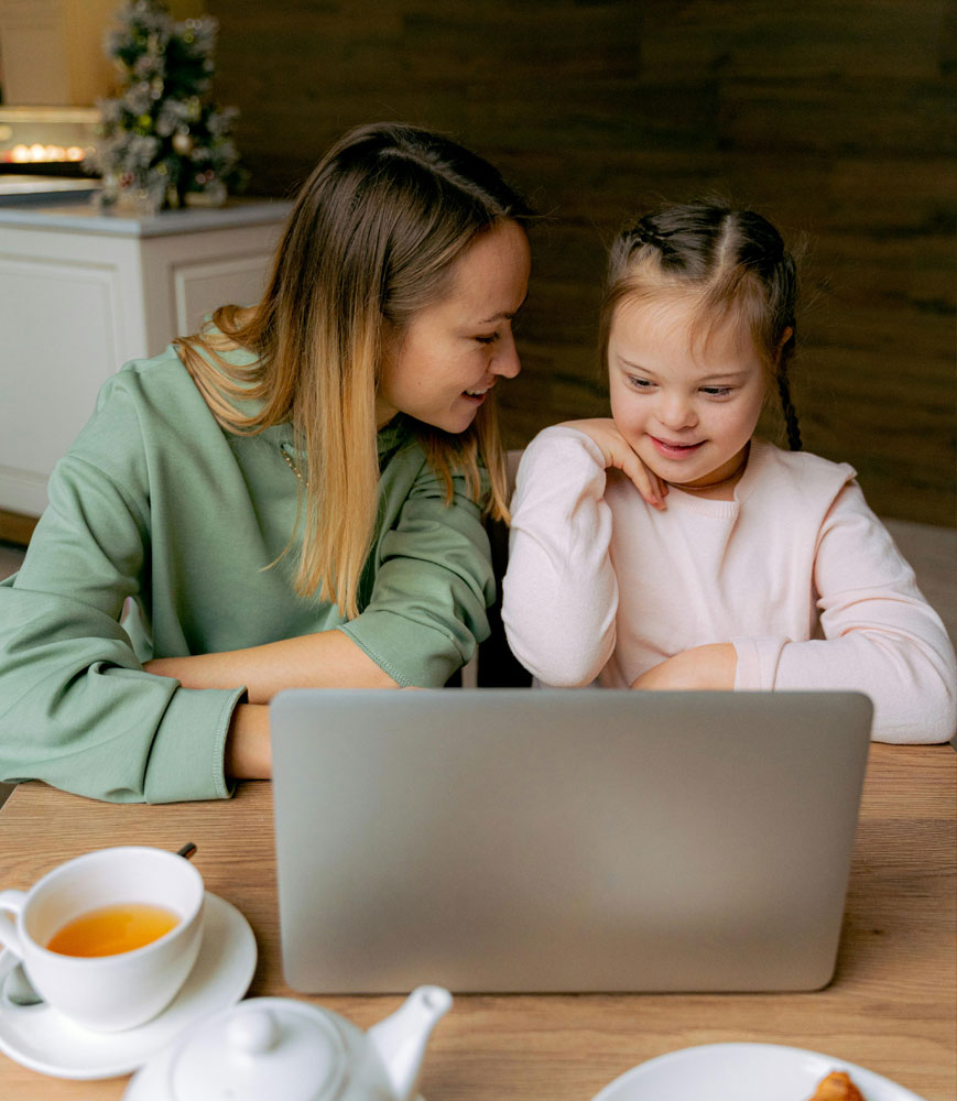 caregiver and young girl with down syndrome look at a laptop while having tea